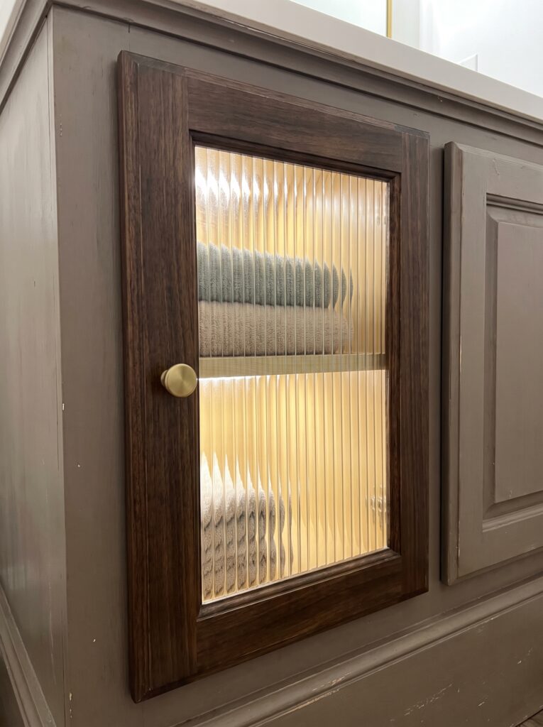 A single wood cabinet door swapped for a reeded glass panel, backlit warmly to show folded towels stacked inside, with a brass knob and matching gray cabinetry on either side