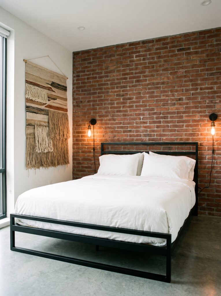 Exposed red brick accent wall behind a black metal platform bed with white bedding and Edison bulb sconces