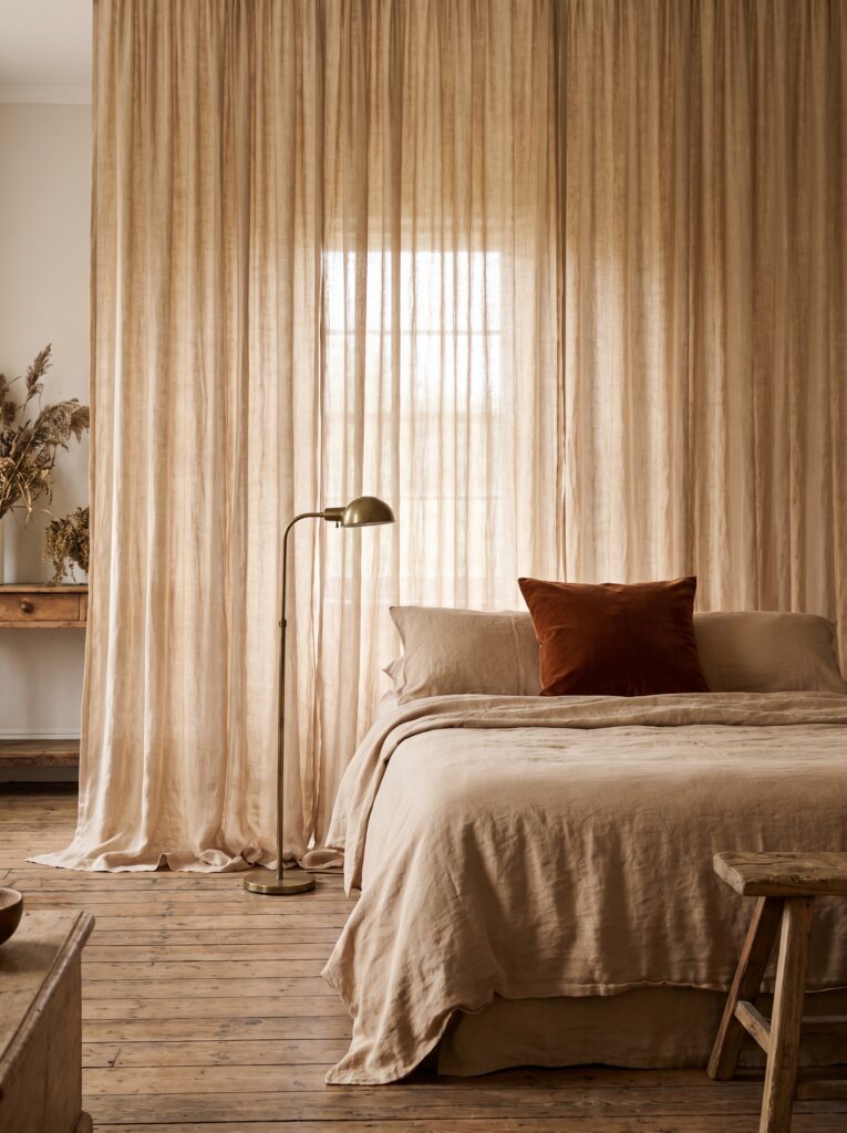 Sheer linen drapery hung floor to ceiling behind a bed in place of a headboard, backlit by natural daylight