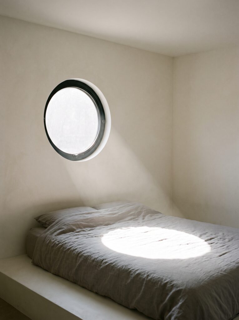 Round porthole-style window framed in matte black metal above a low platform bed, casting a circular beam of light