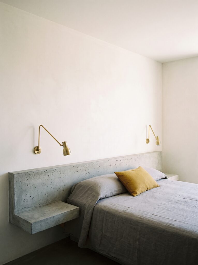  Floating concrete nightstand ledge spanning both sides of a bed with wall-mounted brass reading lights