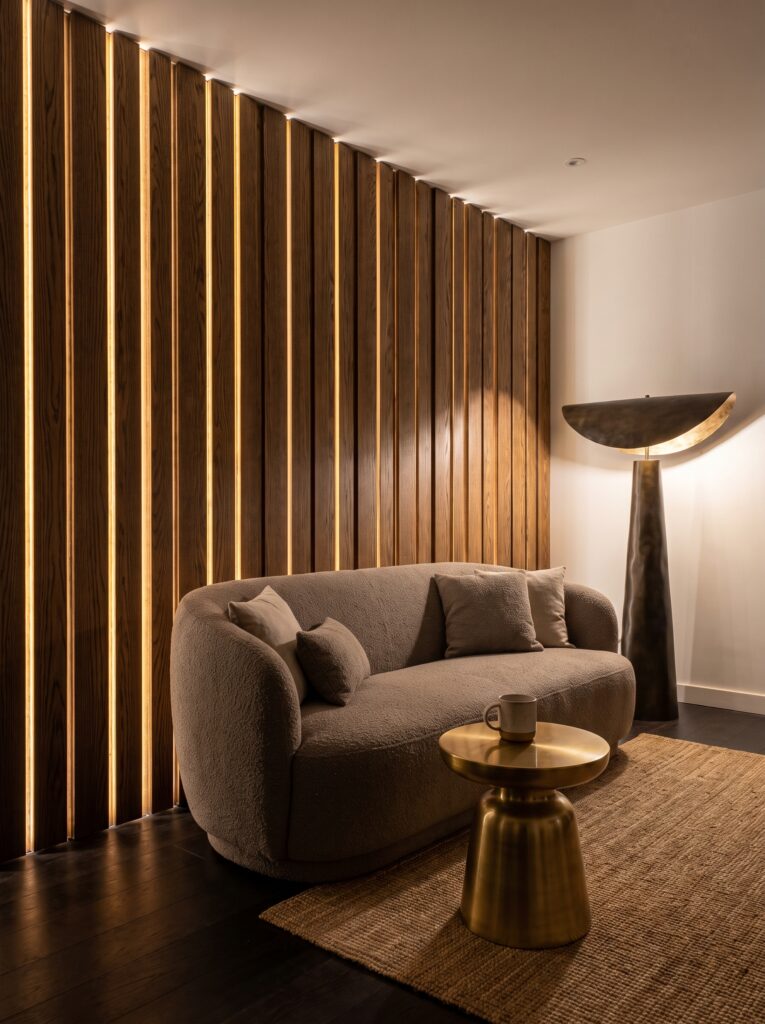 A taupe boucle sofa against a wall of vertical wood slats, each edge lit with warm LED strips, next to a sculptural bronze floor lamp
