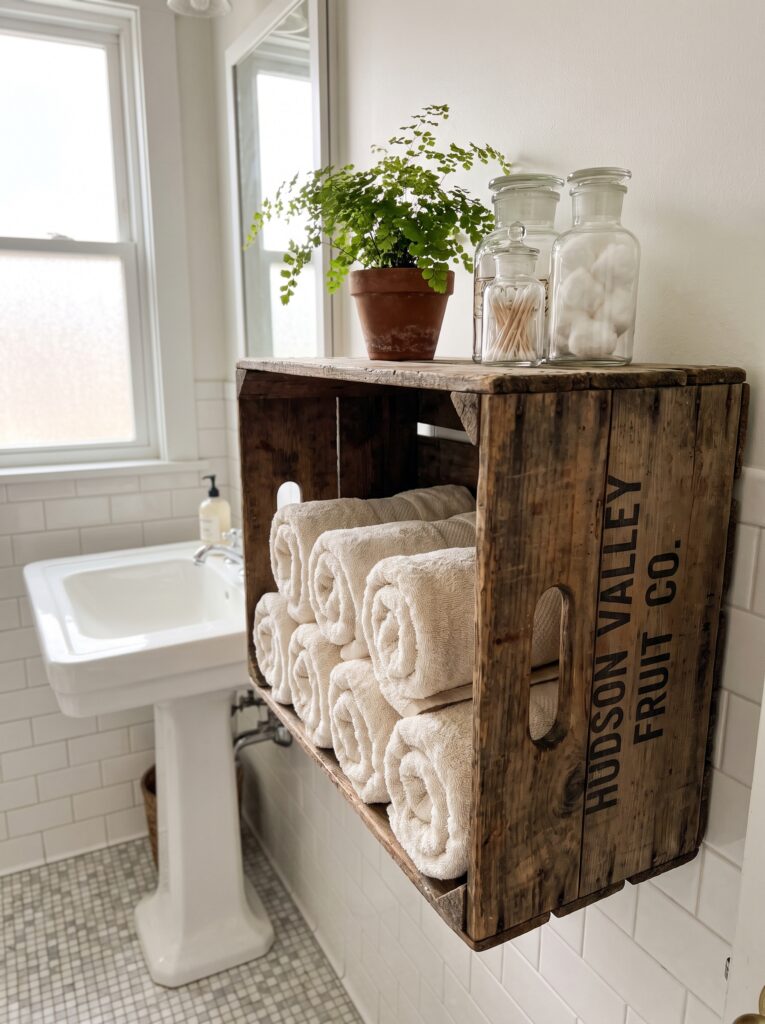 A wooden crate stenciled with vintage lettering, mounted on a bathroom wall and holding rolled towels, a small fern, and glass apothecary jars