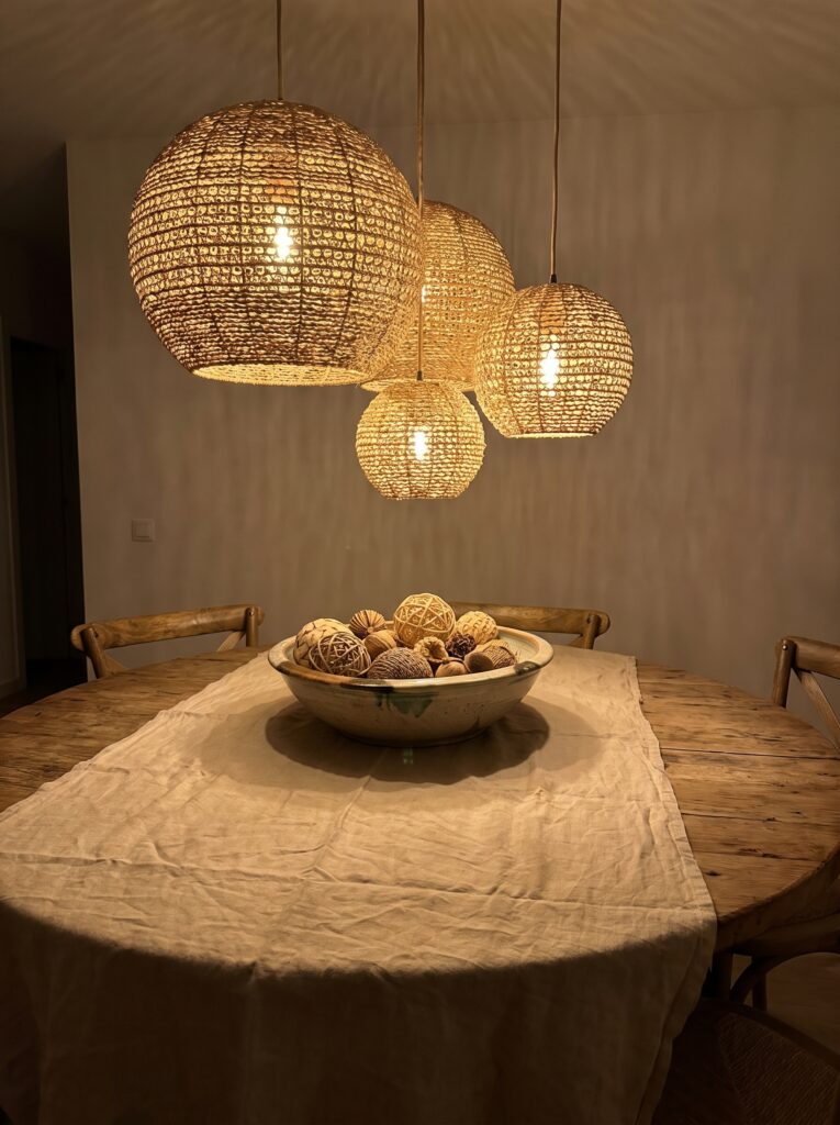 three woven rattan pendant lamps at staggered heights above a round wooden dining table with a linen runner and a bowl of decorative wood spheres