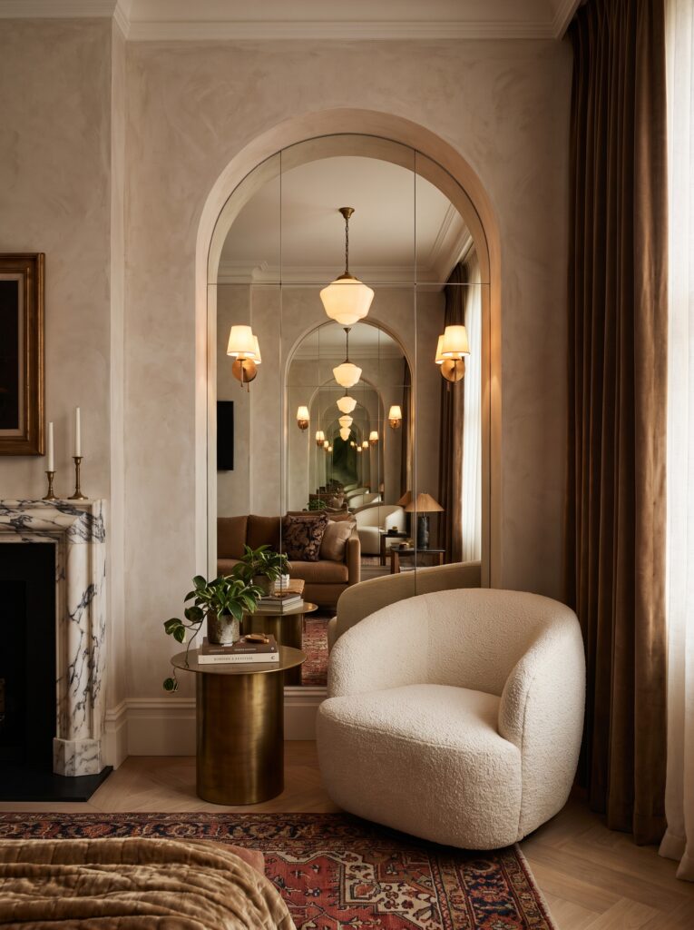 A tall arched mirror set into a plaster wall, reflecting a room full of pendant lights, with a cream boucle chair and brass side table nearby