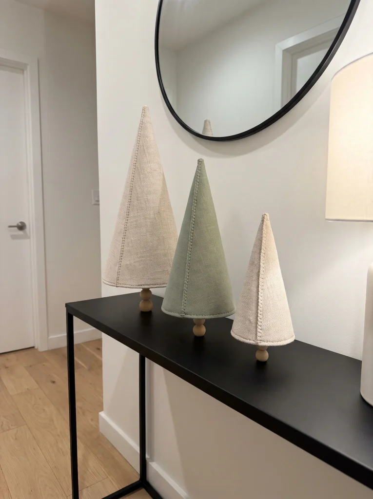 Three linen cone trees in oatmeal, sage and cream on wooden bead stands atop a black console